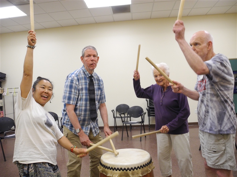 Four people stand around a large drum with drumsticks in their hands
