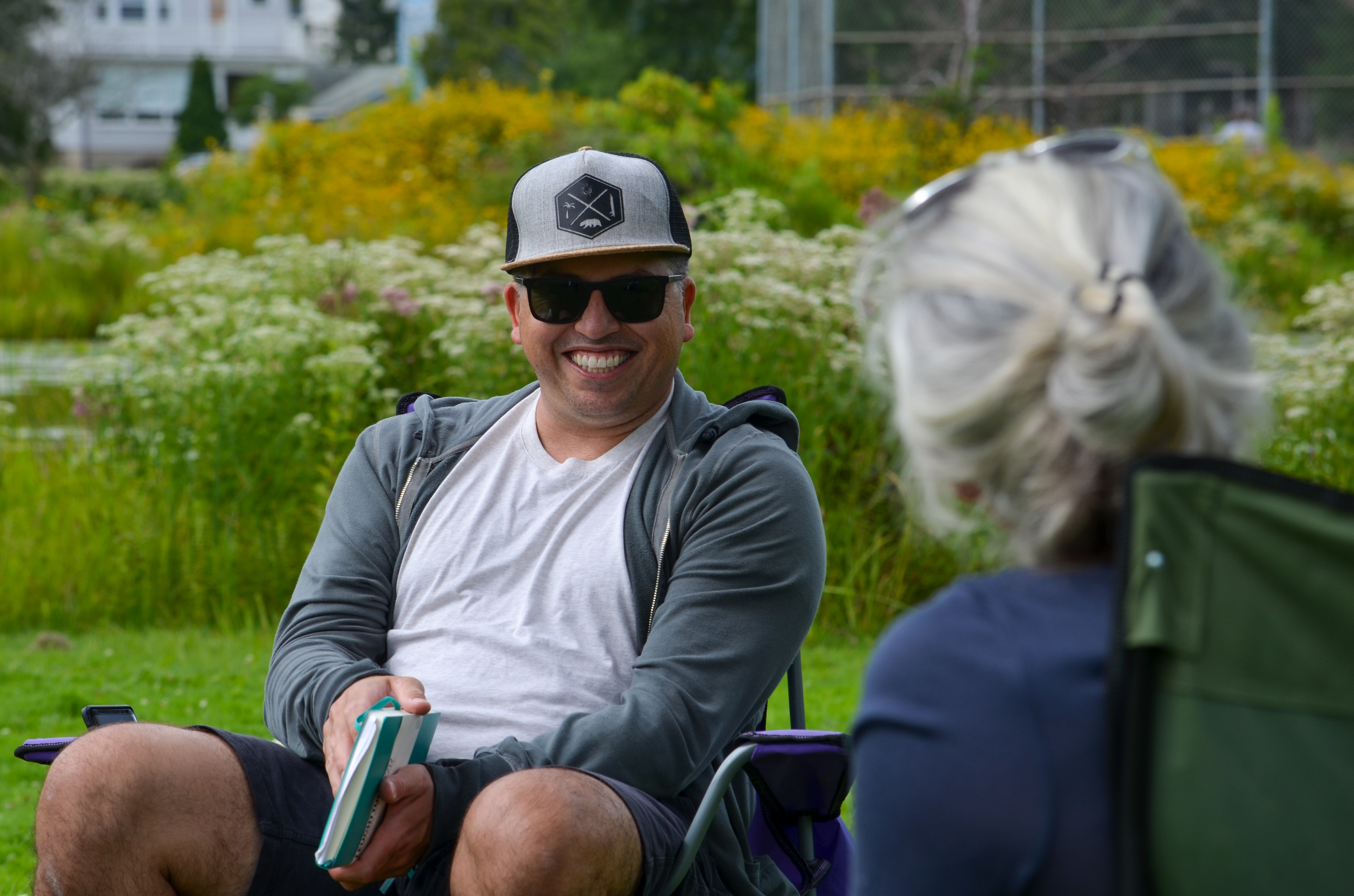 A man in a baseball cap and dark sunglasses sits in a park while smiling at a person in the foreground