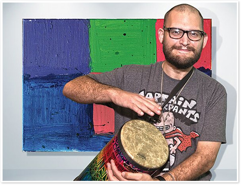 A man wearing glasses plays a drum with a colorful painting behind him