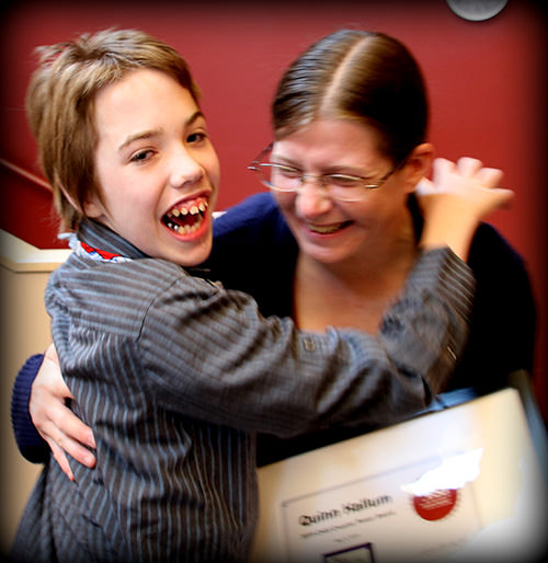 A boy looks back at the camera, smiling, while hugging a woman wearing glasses