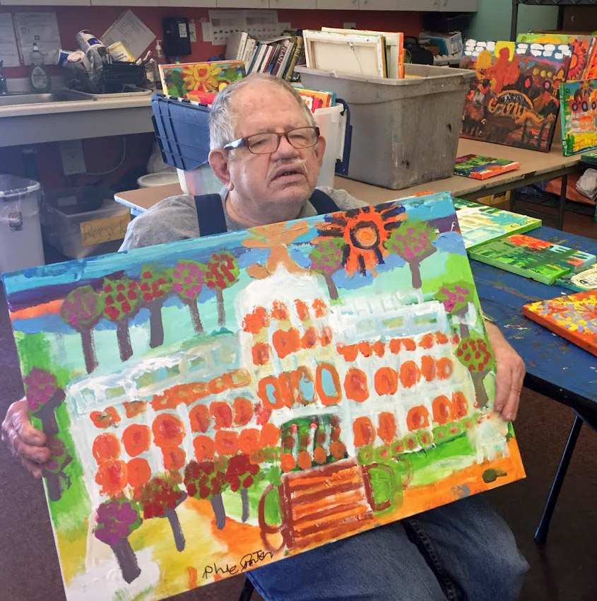 Phil Porter sitting in a chair holding one of his paintings of the capitol building.